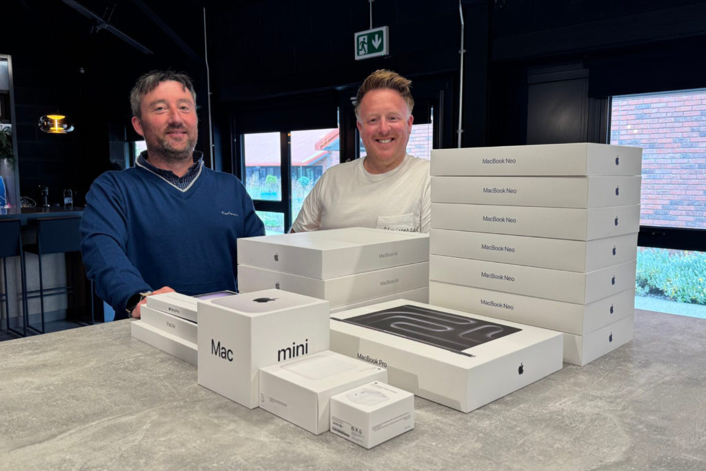 Two smiling business professionals standing behind a large stack of brand-new, boxed Apple computers and devices, celebrating their updated secure hardware infrastructure.